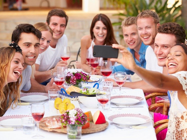 Junge Menschen sitzen draußen im Sommer um einen gedeckten Tisch und machen ein Selfie