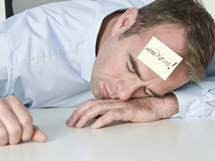 Mann liegt müde mit dem Kopf auf dem Schreibtisch. Auf seiner Stirn klebt ein Post-it "Arbeitslos!"