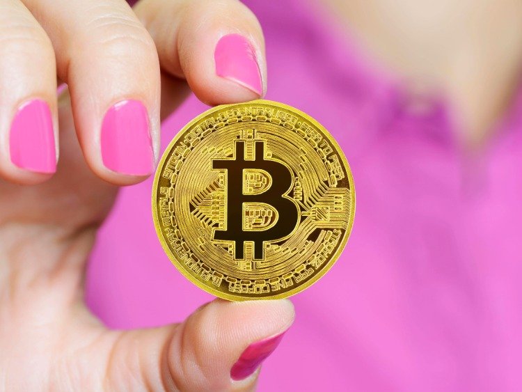 Frau hält zwischen ihren rosa lakierten Fingernägeln einen Bitcoin fest