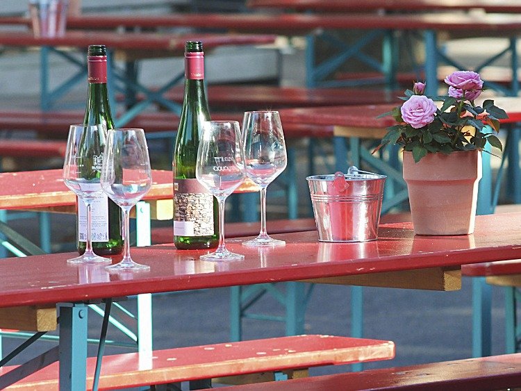 In einem Gartenlokal mit Biertischen sind zwei Weinflaschen zu sehen und vier leere Weingläser