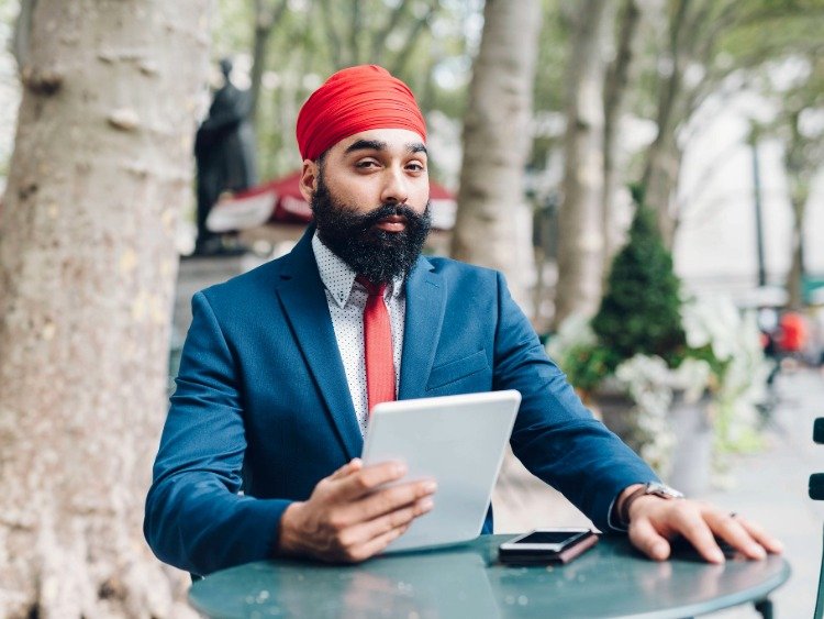 Ein vollbärtiger Mann mit rotem Turban und Businesskleidung hält ein Tablet in der Hand und schaut in die Kamera 