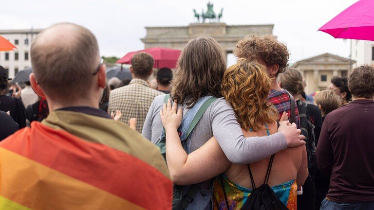 Menschen stehen in einer Demonstration vor dem Brandenburger Tor. Zwei Frauen halten sich im Arm. Ein Mann hat eine Regenbogenfahne um die Schultern gebunden.
