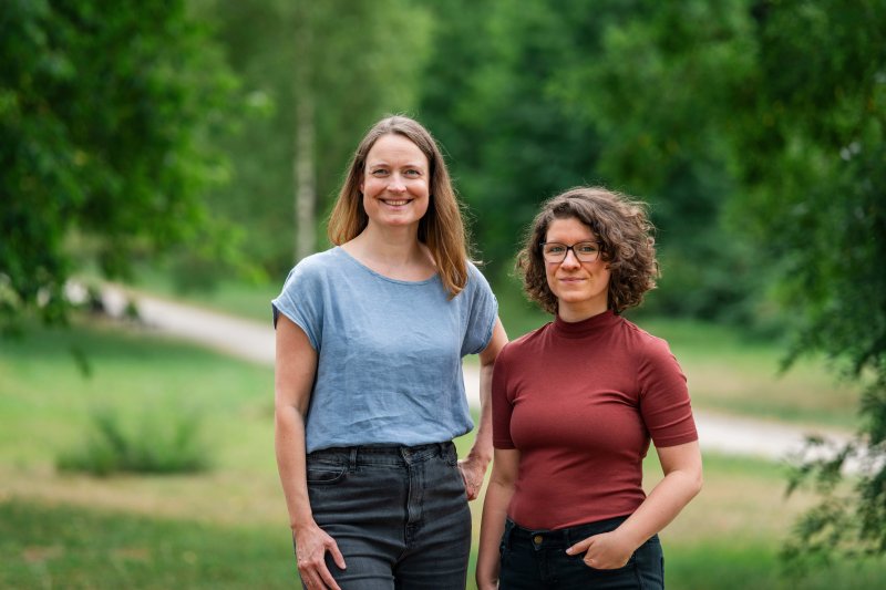 Das Bild zeigt ein Portrait von Ann-Kathrin Büüsker und Ann-Kathrin Horn, den beiden Podcast-Hosts von "Die Klimafrage". Sie stehen nebeneinander in einer grünen Parklandschaft. Im Hintergrund sind Bäume, Wiesen und ein Weg zu sehen. Die Aufnahme zeigt die Personen von etwa der Hüfte aufwärts im Freien.