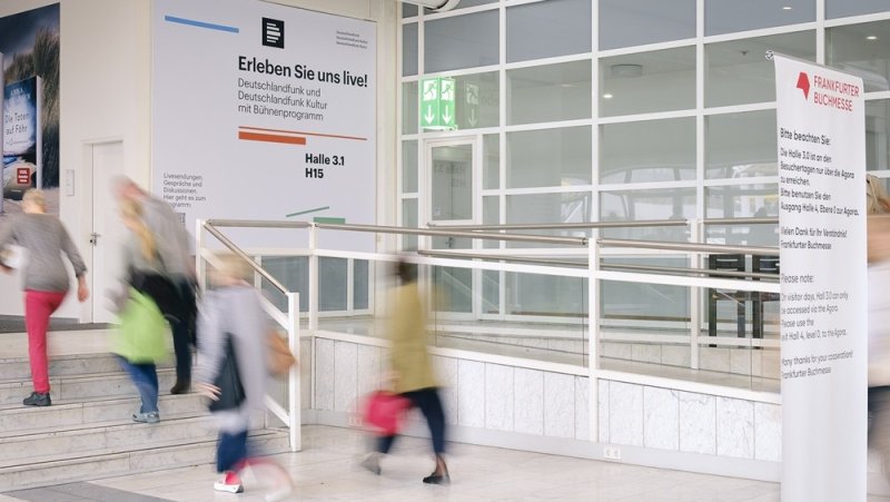 Personen bewegen sich durch den Eingangsbereich einer Messe oder Veranstaltungshalle. An den Wänden hängen Informations- und Veranstaltungsbanner, daneben sind Treppen, eine Rampe und große Fensterflächen zu sehen.
