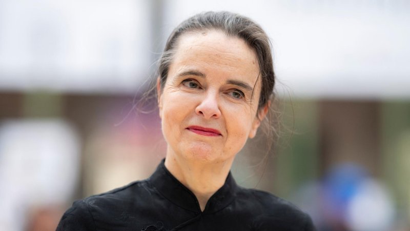 Eine mittelalte Frau mit grau meliertem, zusammengebundenem Haar neigt den Kopf und lächelt leicht.