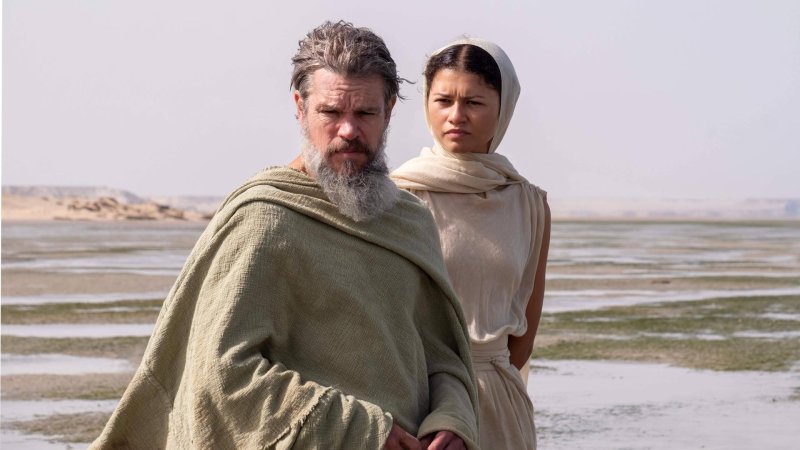 Matt Damon als Odysseus und Zendaya als Athena stehen vor einer Wüsten-ähnlichen Landschaft.