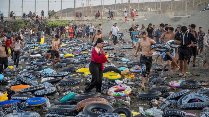 Zahlreiche junge Menschen an einem Strand, der übersät ist mit Plastikschwimmreifen. Im Hintergrund ein hoher Zaun. 