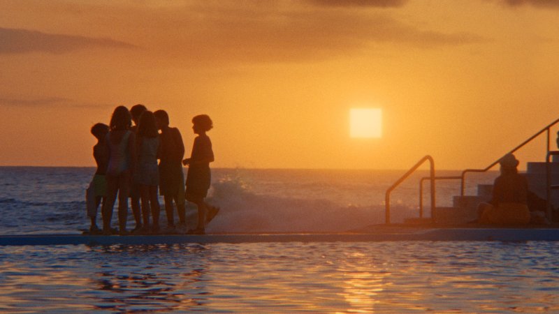 Eine Gruppe Kinder steht am Meer, im Hintergrund strahlt goldgelb der Sonnenuntergang. Die Sonne ist quadratisch.