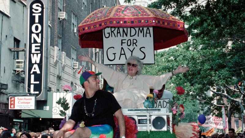Eine ältere Frau auf einem Paradewagen, unter einem Schirm. Hinter sich hat sie ein Schild: "Grandmas for gays"
