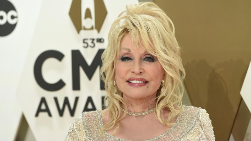 Die Countrymusikerin Dolly Parton steht bei einer Preisverleihung vor einer Wand und lächelt in die Kamera. Sie träg ein gold-glitzerndes Oberteil. 