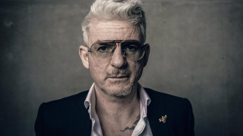 Der Schriftsteller Heinz Strunk in Hemd und Sakko und mit getönter Brille steht vor grauem Hintergrund. Er blickt in die Kamera. 