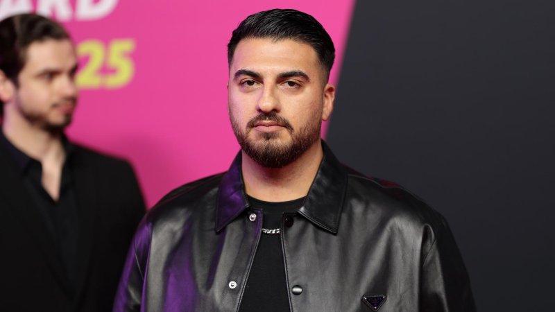 Tahsim Durgun steht auf einer Bühne vor einem pinkfarbenen Hintergrund, blickt direkt in die Kamera und trägt eine schwarze Jacke.