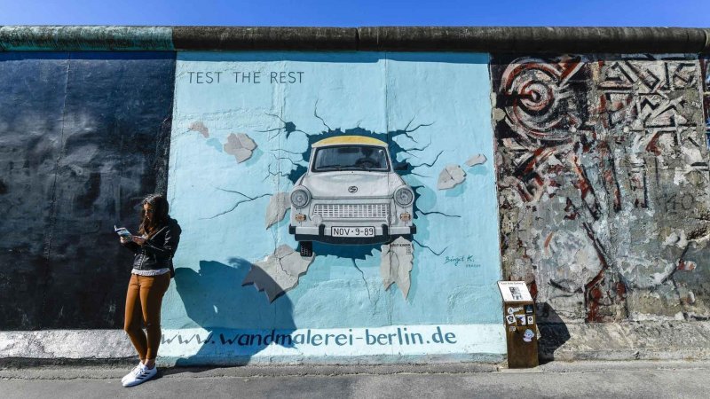 eine Frau an der Berliner East Side Galery vor der Berliner Mauer und liest in einer Broschüre. 