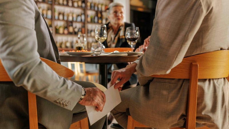 Ein weißes Kuvert geht an einem Restauranttisch von einer Hand zur anderen