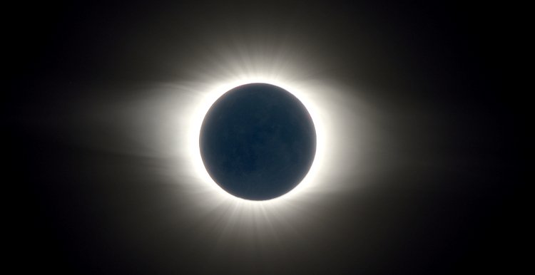 Der pechschwarze Kreis des Neumonds ist von einem weißlichen Lichtkranz umgeben, der an vielen Stellen linienförmige Strukturen zeigt.
