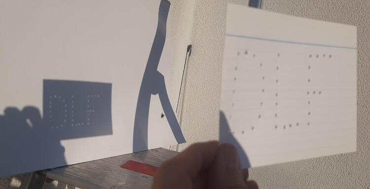 Eine Hand hält ein Stück Papier, in das Löcher gestanzt sind, die den Schriftzug DLF ergeben. Ein Stück dahinter ist der Schatten des Blattes zu sehen, in dem perfekte Kreise aus Sonnenlicht DLF ergeben.