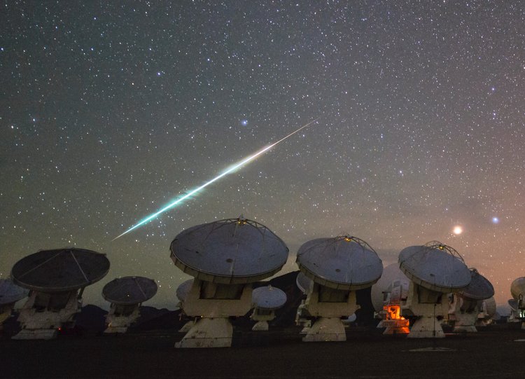 In einer Wüstenlandschaft stehen mehrere Radioteleskope, die wie riesige Satellitenschüsseln aussehen. Über den gestirnten Himmel darüber zieht eine sehr helle Sternschnuppe