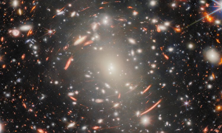 Um einen Galaxienhaufen unterschiedlich großer, meist ovaler weißer Objekte sind viele rötliche Strukturen zu sehen. Die meisten sind in die Länge gezogen und zum Zentrum des Haufens hin gebogen.